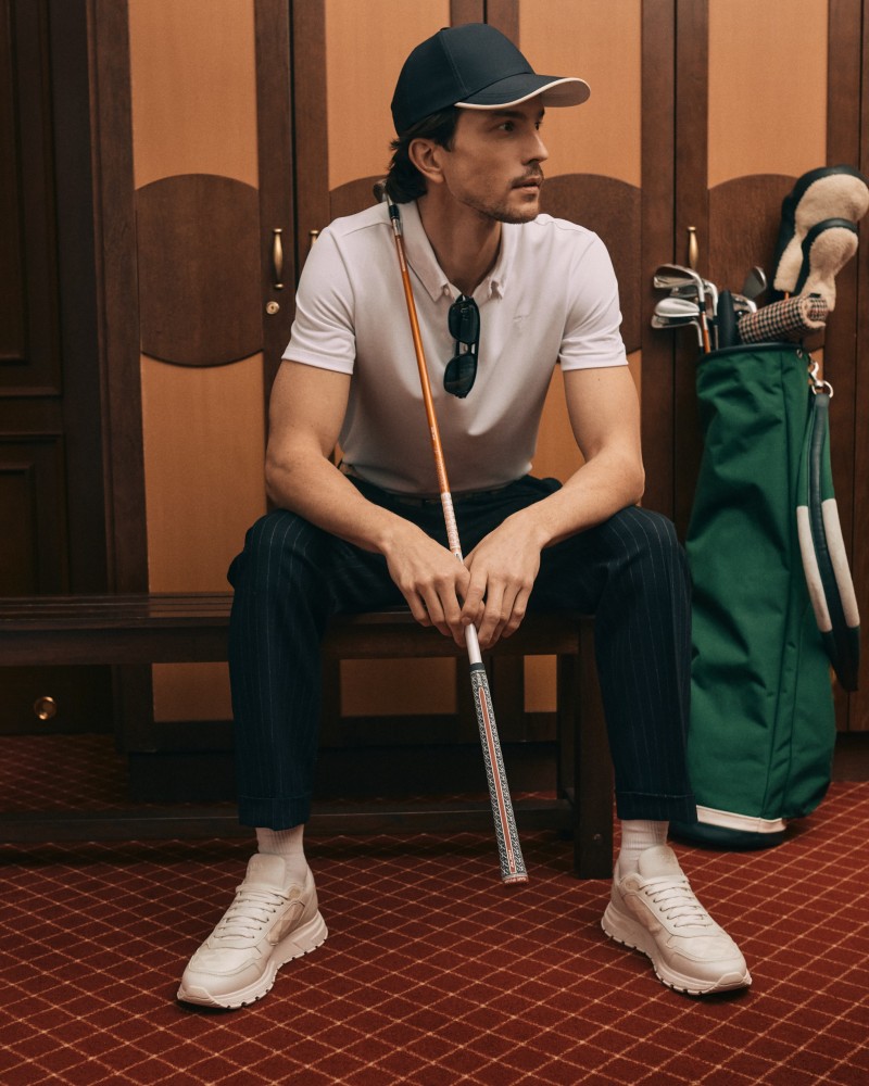 Mężczyzna w białym polo i spodniach w prążki, siedzący na ławce w szatni obok torby golfowej