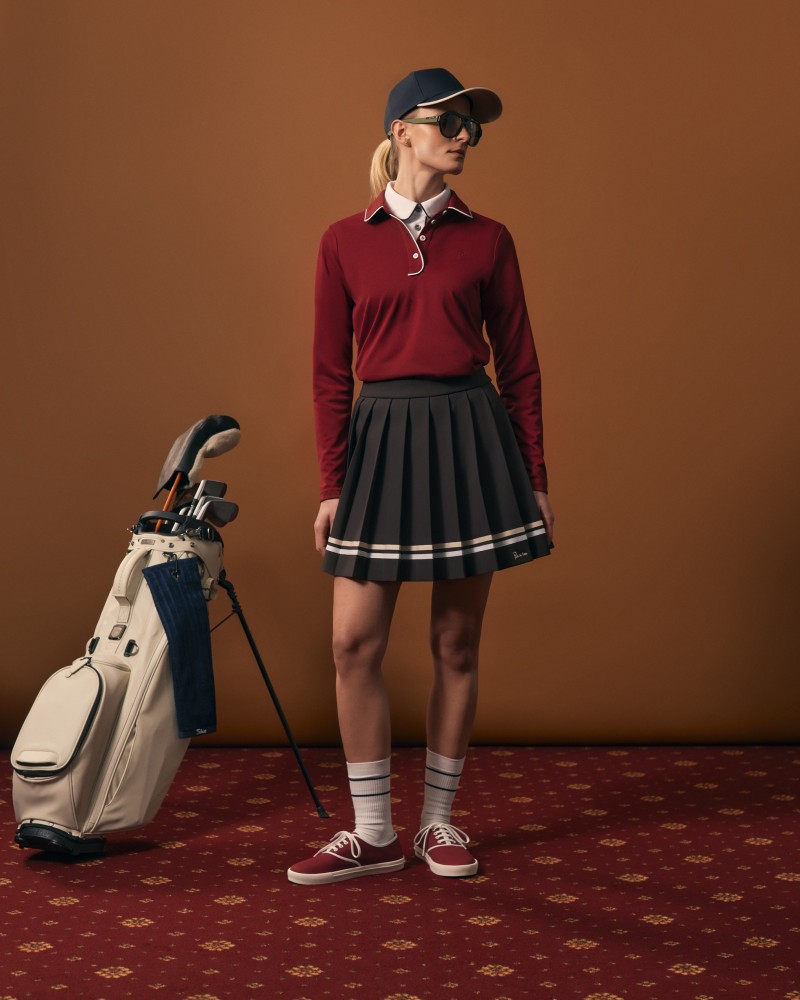 Modelka w bordowym polo i czarnej plisowanej spódniczce, obok torby golfowej