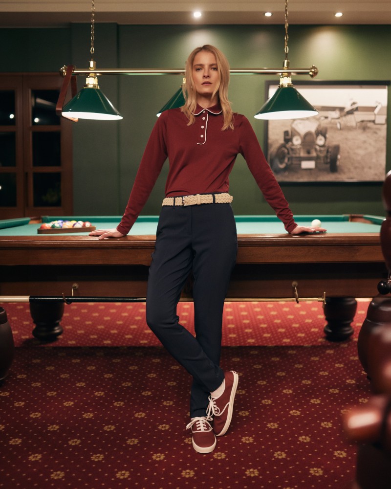 Modelka w bordowym polo, granatowych spodniach i plecionym pasku, przy stole bilardowym