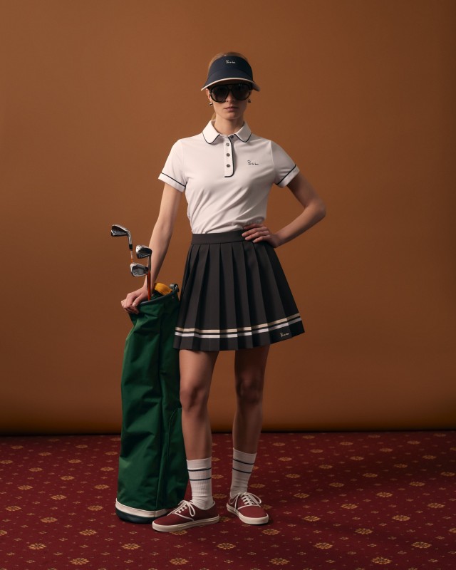 Modelka w białym polo i czekoladowej plisowanej spódniczce, w okularach, z torbą golfową